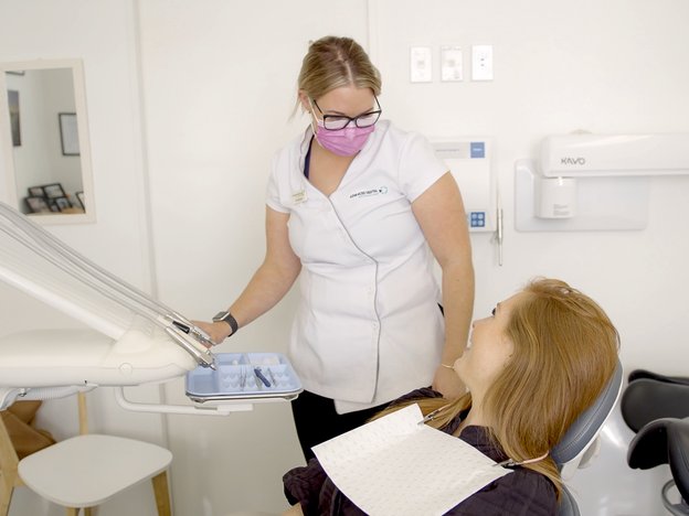 A female in a white uniform, pink mask and black rimmed glasses assists a woman with red hair lying down in a dental chair in a dentist's office.