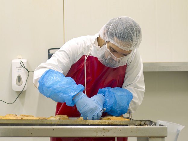 A female in a white jacket, red apron and blue gloves assembles meat on a tray in a factory. She is wearing a white hairnet and facemask for protection.