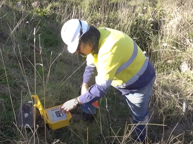 A man wearing a fluorescent yellow and navy-blue jacket and white helmet stands in a grassy field.
