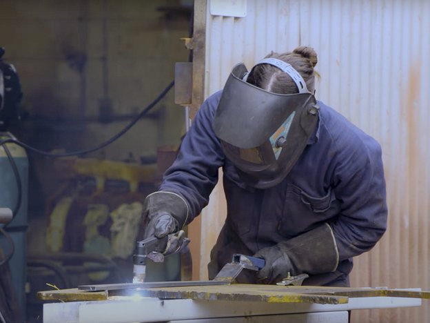 A female wearing a blue jumpsuit with a protective mask and black gloves welds a piece of metal on a work bench. She is in a factory and is using a welding gun.