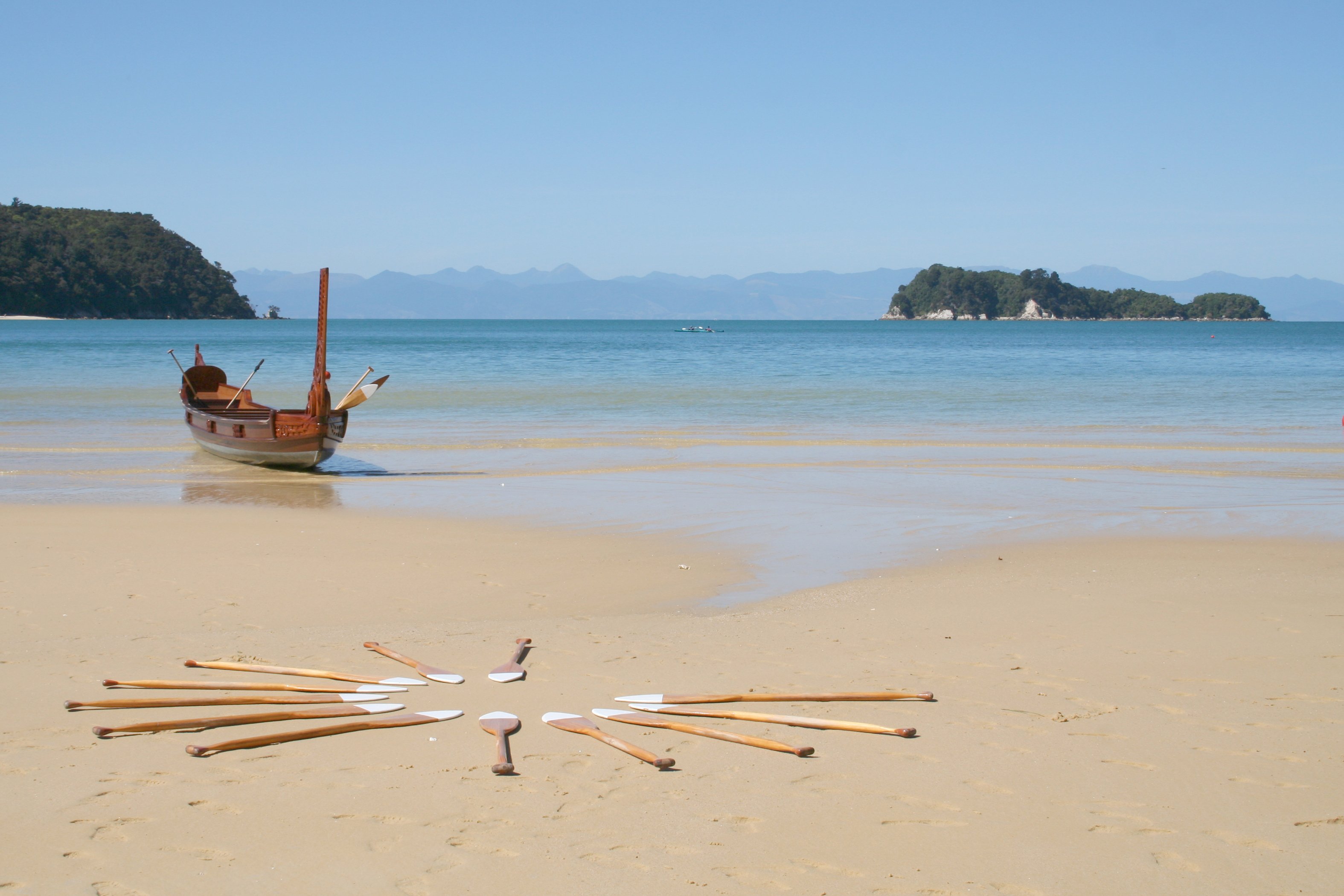 A waka is pulled onto shore with oars laid in a circle on the sand.