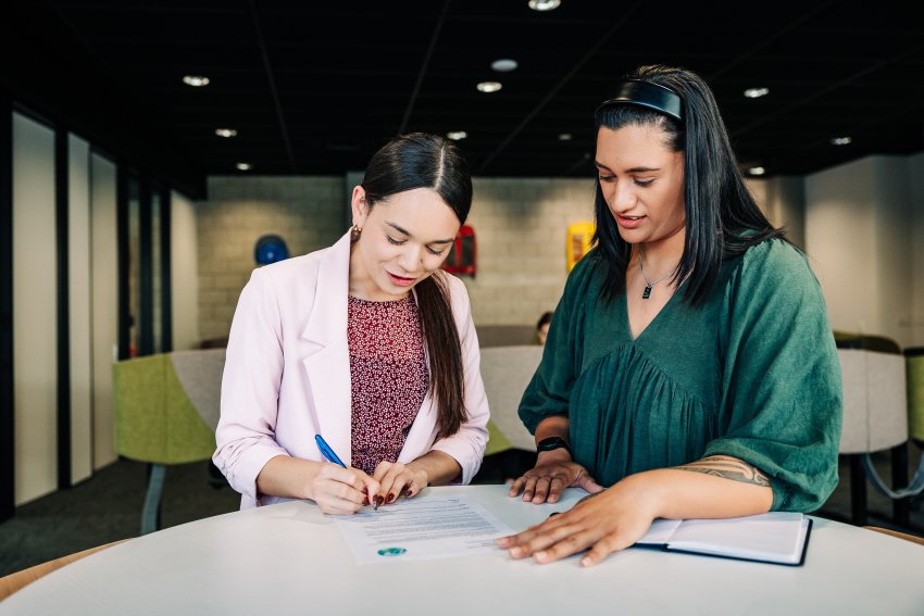 Two people look at a document on a table, one is filling it out with a pen.