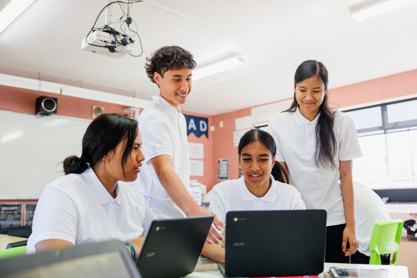Four students look at a laptop. They are in a classroom, wearing a school uniform.