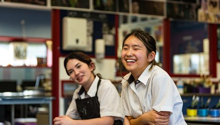 Two girls smile in art class, while sitting at their desk. Behind them are painting tools and photography on the wall.