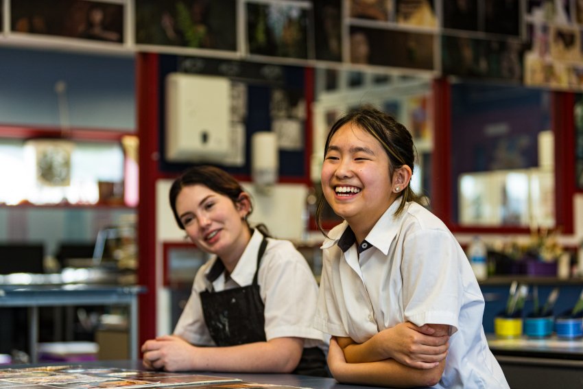 Two girls smile in art class, while sitting at their desk. Behind them are painting tools and photography on the wall.