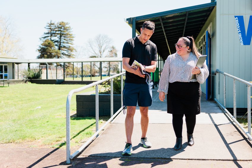 Two people look at a workbook. They are walking down a ramp, outside a school classroom.