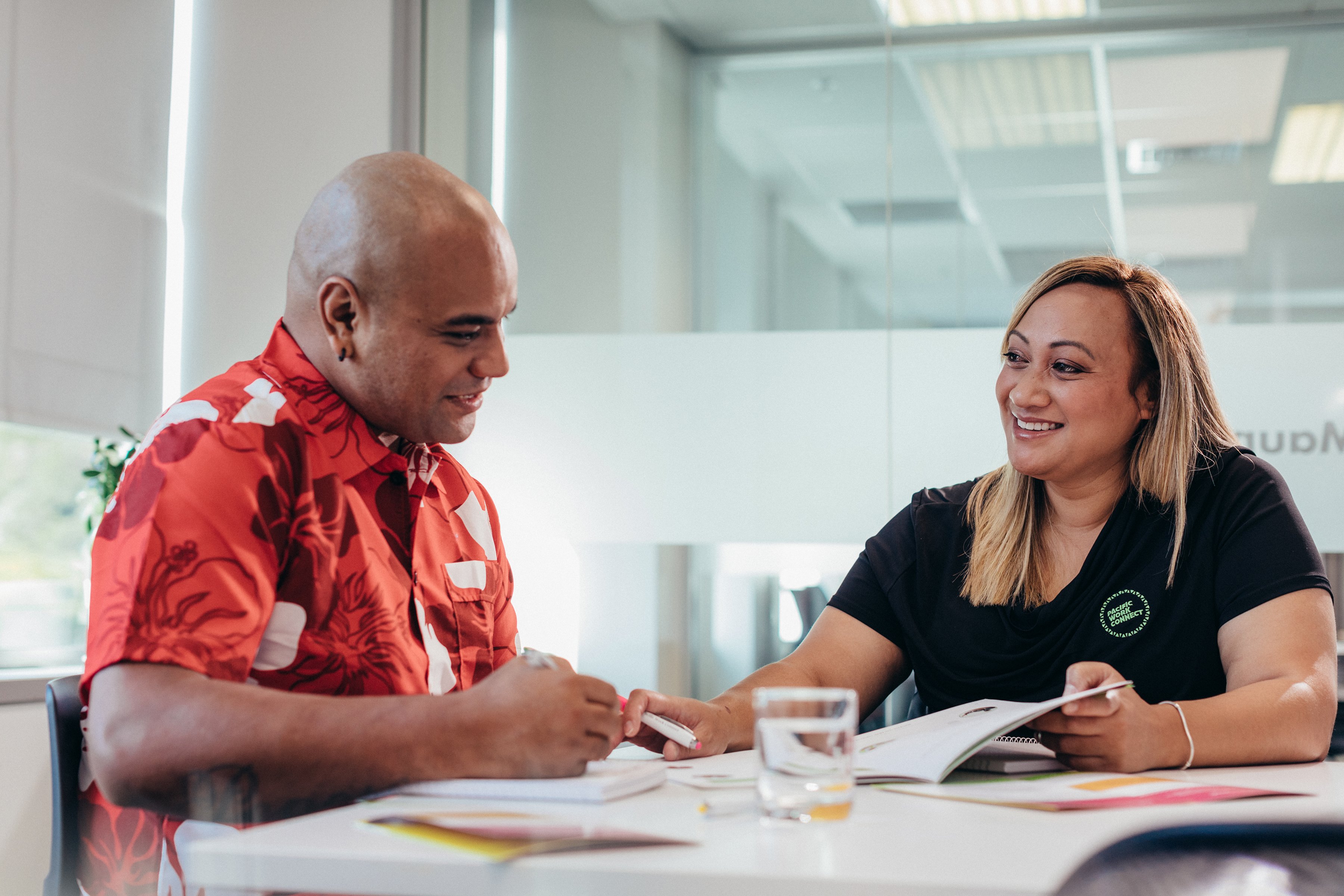 Two people sit at a table, looking at a workbook. One is showing a page to the other, they both look happy.