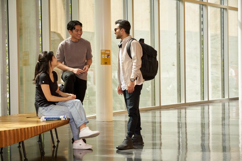 Three students talk in a large indoor space. One is sitting on an antique wooden chair while the others are standing.