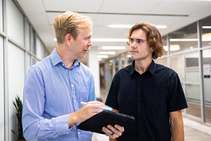 An office worker talk to a new employee. One worker is holding a pen and a tablet device.