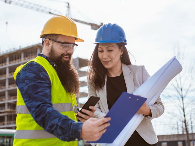 Male wearing a high-viz vest. Female wearing a jacket. Both are wearing hard hats. They're standing in front of a construction site and they're holding a phone, clipboard and an architectural plan. There's a crane in the background.