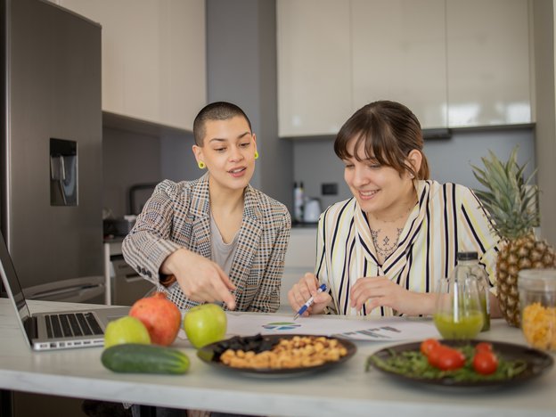 Two people sit at a kitchen bench studying food. In front of them are fruit, vegetables and nuts.