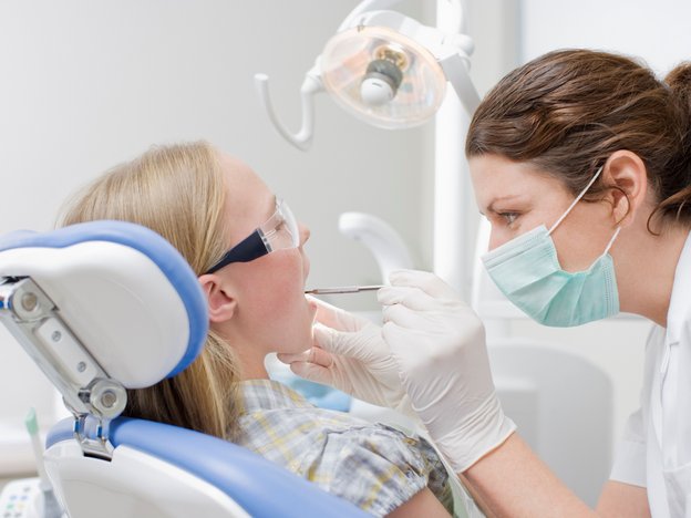 Oral health therapist, wearing a face mask and gloves, examining a young females' mouth who is sitting in a dentist's chair.
