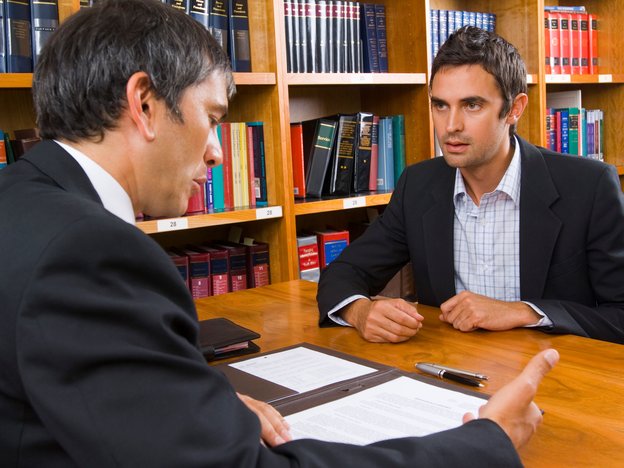 wo men sitting opposite each other with a desk between them. One man is looking down at some documents.