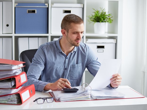 A person sits at their work desk reading a piece of paper and using a calculator.