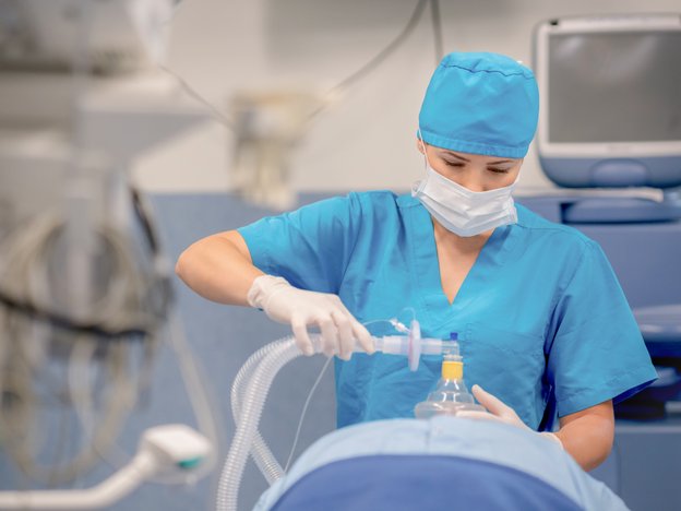 A female anaesthetist in blue medical scrubs places anaesthesia mask on patients face.