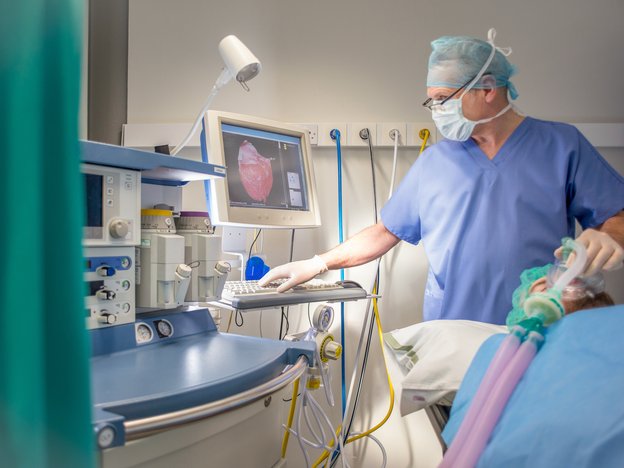 A male anaesthetist in blue medical scrubs observes a computer while a patient is under general anaesthetic. An organ is displayed on the screen.