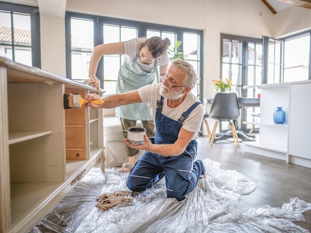 An older man and woman apply finish to a wooden bench, inside a modern home.