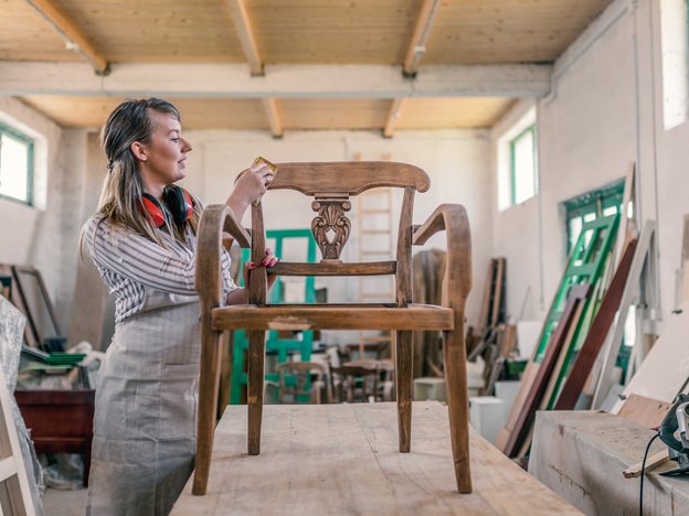 A woman applies finish to a wooden chair that stands on a table. She stands in a workshop and is surrounded by other wooden pieces.
